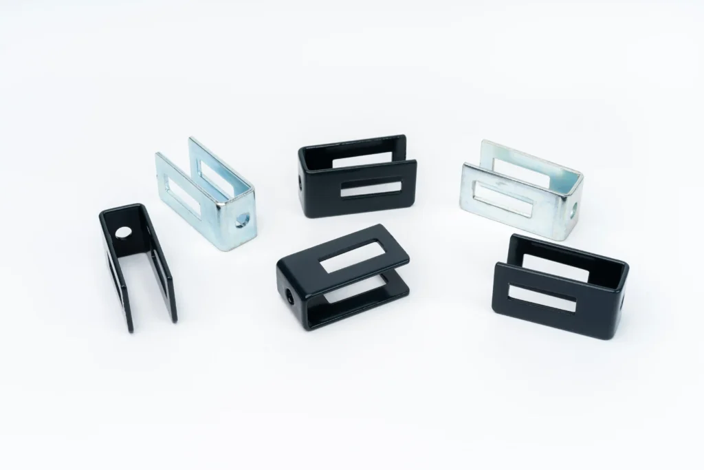 Uniwersalna klamra do montażu ogrodzeń – metalowe uchwyty montażowe w różnych wariantach kolorystycznych (czarne i ocynkowane), ułożone na białym tle.